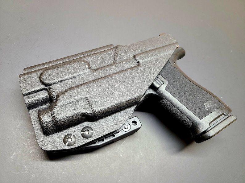 May include: A black polymer holster with a textured surface and a black handgun inside. The holster has a belt clip and three screws on the bottom.