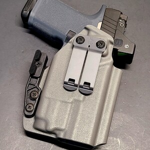 May include: A gray handgun with a black slide and a red dot sight is placed inside a gray holster. The holster has two metal clips and a black attachment. The background is a dark gray surface.