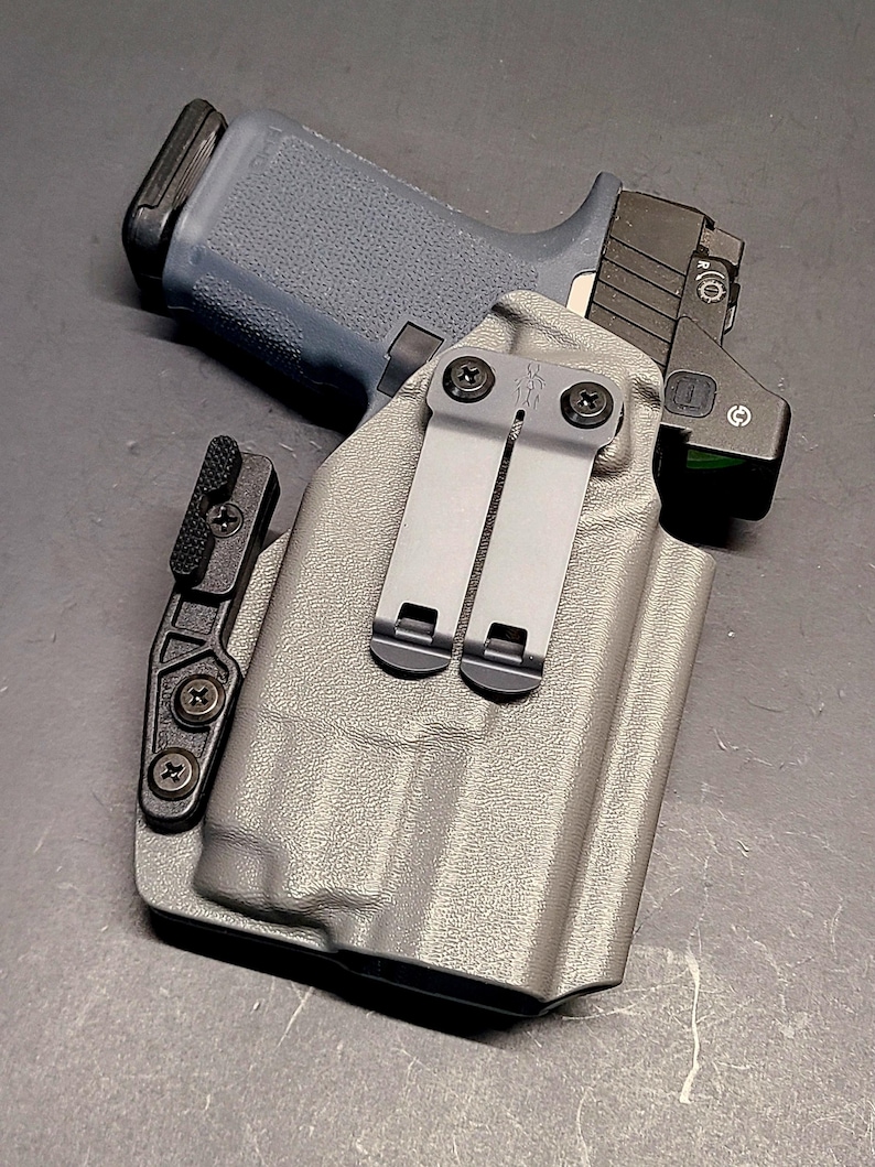 May include: A gray handgun with a black slide and a red dot sight is placed inside a gray holster. The holster has two metal clips and a black attachment. The background is a dark gray surface.