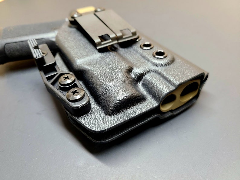May include: A black polymer holster with a tan leather lining and a metal clip. The holster has two screw holes on the side and two holes on the top.