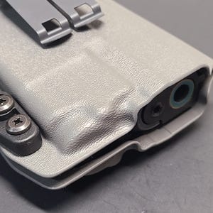 May include: A light gray, textured polymer holster with a black attachment clip and silver screws. The holster has a molded design to fit a firearm, with the barrel end visible. The holster features a gray metal clip.
