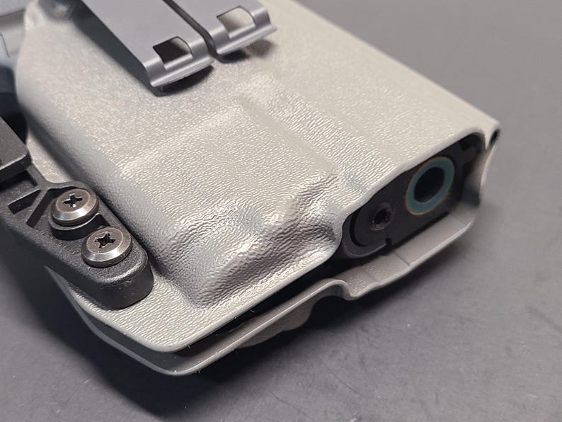 May include: A light gray, textured polymer holster with a black attachment clip and silver screws. The holster has a molded design to fit a firearm, with the barrel end visible. The holster features a gray metal clip.
