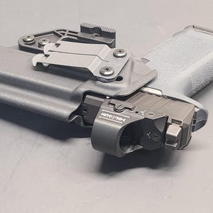 May include: A grey handgun in a black holster with a mounted light. The holster features a belt clip and is secured with screws. The light has the text "HOLOSUN" printed on it. The handgun rests on a grey surface.