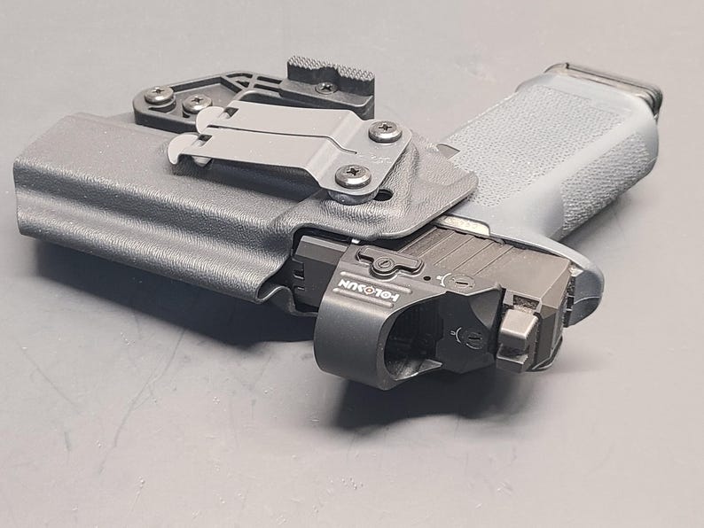 May include: A grey handgun in a black holster with a mounted light. The holster features a belt clip and is secured with screws. The light has the text "HOLOSUN" printed on it. The handgun rests on a grey surface.