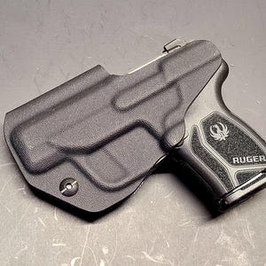 May include: A black Ruger handgun in a black holster. The handgun has a textured grip with the Ruger logo and the word "RUGER" printed on it. The holster is molded to the shape of the gun.