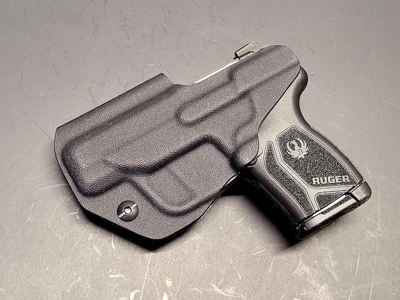 May include: A black Ruger handgun in a black holster. The handgun has a textured grip with the Ruger logo and the word "RUGER" printed on it. The holster is molded to the shape of the gun.