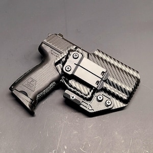 May include: A black handgun with a textured grip and the marking "P 2000" is shown next to a black and gray carbon fiber pattern holster. The holster has a metal clip and is secured with screws.