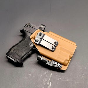 May include: A black handgun with a red dot sight is placed next to a tan holster. The holster has a metal clip and a black attachment. The items are on a dark surface.