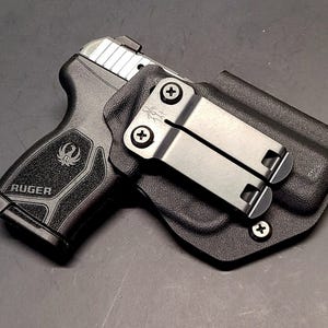 May include: Black Ruger handgun in a black holster. The handgun has a silver slide and the Ruger logo is visible on the grip. The holster has a metal clip for attachment.
