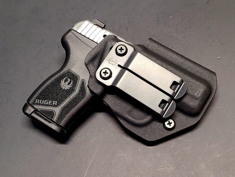 May include: Black Ruger handgun in a black holster. The handgun has a silver slide and the Ruger logo is visible on the grip. The holster has a metal clip for attachment.
