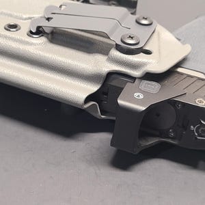 May include: A close-up of a gray pistol in a light gray holster. The pistol has a black attachment with a small circular opening. The holster has a metal clip and is secured with screws.