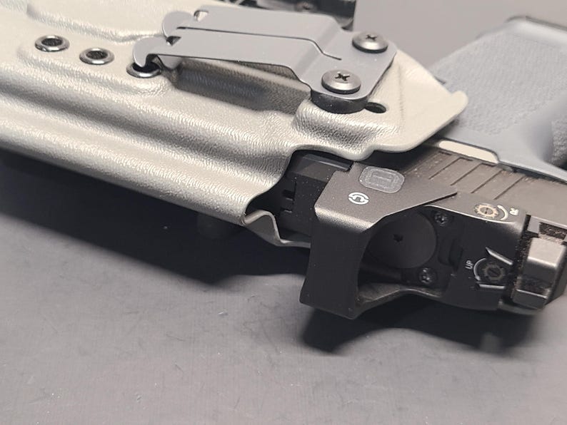 May include: A close-up of a gray pistol in a light gray holster. The pistol has a black attachment with a small circular opening. The holster has a metal clip and is secured with screws.