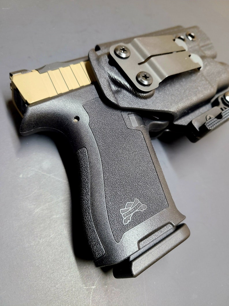 May include: A black handgun with a tan slide and a black holster. The holster has a metal clip and a black strap. The handgun has a black grip with a logo that says "Palmetto State Armory".