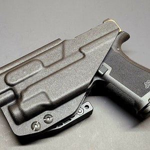 May include: A black polymer holster with a black handgun inside. The holster has a textured surface and a clip on the side. The handgun has a black polymer frame and a black metal slide.