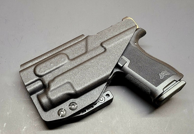 May include: A black polymer holster with a black handgun inside. The holster has a textured surface and a clip on the side. The handgun has a black polymer frame and a black metal slide.