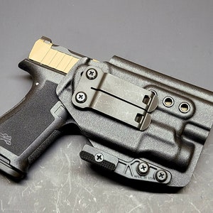 May include: A black polymer holster with a black metal clip and a black handgun inside. The holster has a "MADE IN USA" label on the bottom.