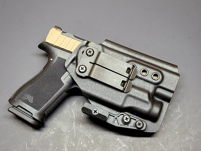 May include: A black polymer holster with a black metal clip and a black handgun inside. The holster has a "MADE IN USA" label on the bottom.