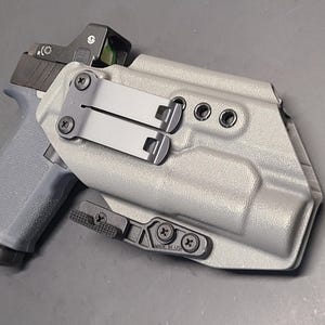 May include: A gray pistol with a red dot sight is partially inserted into a gray holster. The holster has black metal clips and screws. The pistol grip is visible, and the overall design suggests a focus on firearm accessories.