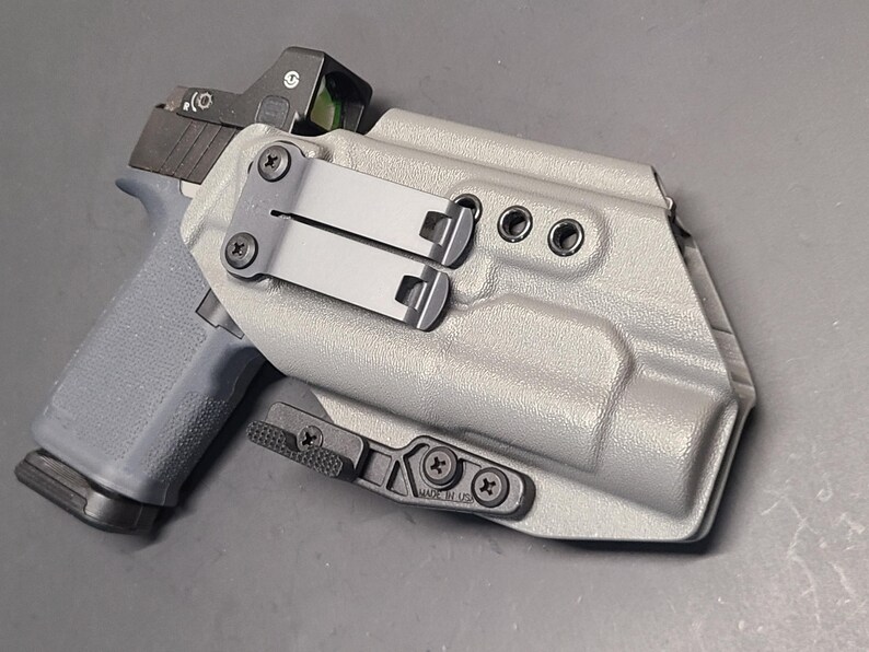 May include: A gray pistol with a red dot sight is partially inserted into a gray holster. The holster has black metal clips and screws. The pistol grip is visible, and the overall design suggests a focus on firearm accessories.