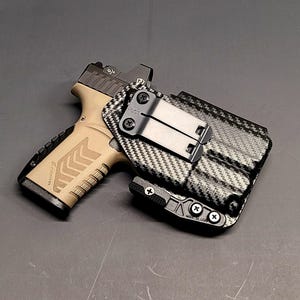 May include: A tan and black handgun with a black holster. The handgun has a tan frame and black slide with a red dot sight. The holster is black with a carbon fiber pattern and a metal clip.