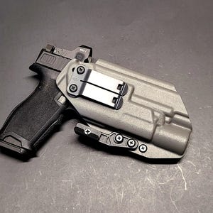 May include: A black handgun with a gray holster and a red dot sight. The holster has a metal clip and is secured with screws. The gun is resting on a dark surface.