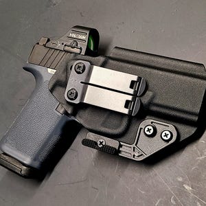 May include: A black handgun with a blue grip and a red dot sight is secured in a black holster. The holster has a metal clip and is mounted with screws. The firearm is positioned on a dark surface.