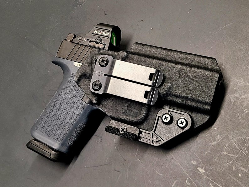 May include: A black handgun with a blue grip and a red dot sight is secured in a black holster. The holster has a metal clip and is mounted with screws. The firearm is positioned on a dark surface.