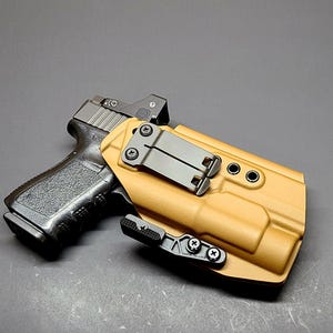 May include: A black handgun with a red dot sight is partially inserted into a tan-colored holster. The holster has a black clip and hardware. The image is set against a dark gray background.