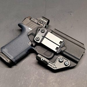 May include: A black handgun with a red dot sight in a black holster. The holster features a metal clip and is secured with screws. The gun has a dark blue grip and a black magazine.