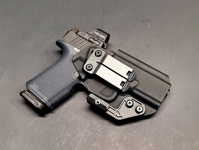 May include: A black handgun with a red dot sight in a black holster. The holster features a metal clip and is secured with screws. The gun has a dark blue grip and a black magazine.