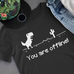 Pode incluir: Camiseta preta com um dinossauro pixelado branco, um cacto e o texto "You are offline!" impresso nela. A camiseta está sobre uma superfície cinza com um chapéu estampado floral e um par de fones de ouvido brancos.