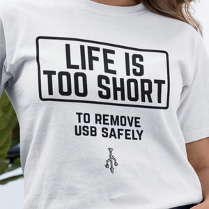 Op de afbeelding: Wit T-shirt met een zwarte afbeelding met de tekst "Het leven is te kort om USB-sticks veilig te verwijderen".