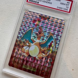 May include: A 1996 Japanese Bandai Charizard-Prism Carddass Vending trading card graded GEM MT 10. The card features a holographic Charizard illustration, the text "MONSTERS COLLECTION," and English descriptive text in the background.