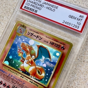 May include: A graded 1998 Japanese Charizard Holo CD Promo card. The card features a vibrant illustration of Charizard, an orange and blue dragon-like creature. The card is encased in a protective case with a label indicating its condition and serial number.