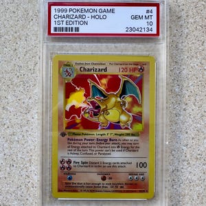 May include: A Charizard Holo 1st Edition Pokémon card from 1999, graded GEM MT 10. The card features a yellow and orange background with a Charizard illustration. The card is encased in a protective holder.