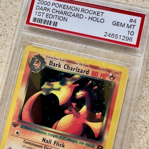 Może przedstawiać: Oceniona karta Pokémon Rocket Dark Charizard Holo 1st Edition z 2000 roku. Karta jest zamknięta w ochronnym etui z czerwoną etykietą z napisem "GEM MT 10". Karta zawiera żywą ilustrację Charizarda.