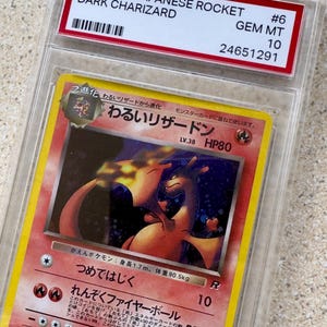 May include: A graded 1999 Japanese Rocket Dark Charizard Pokémon card. The card is encased in a protective case with a red label that reads "1999 P.M. JAPANESE ROCKET DARK CHARIZARD #6 GEM MT 10". The card features a Charizard illustration.