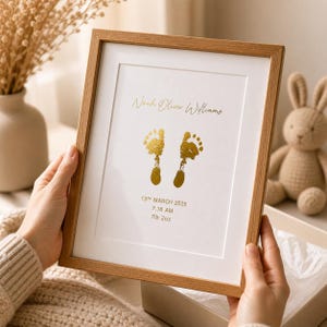 May include: A framed print with a light wood frame. The print features two gold-colored baby footprints, the name "Noah Oliver Williams", and the date, time, and weight of the baby. The frame is held by a person.