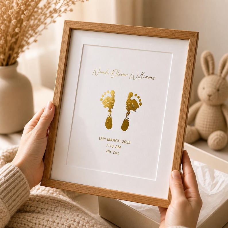 May include: A framed print with a light wood frame. The print features two gold-colored baby footprints, the name "Noah Oliver Williams", and the date, time, and weight of the baby. The frame is held by a person.
