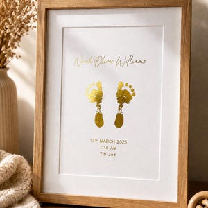 Op de afbeelding: Een ingelijste print met een licht houten frame. De print toont gouden voetstappen, de naam "Noah Oliver Williams" en geboortegegevens: 13 maart 2025, 7:18 uur, en 3,2 kg.