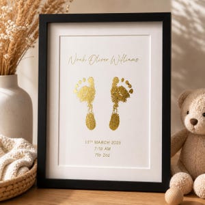 May include: Framed print with gold foil baby footprints. The print includes the name "Noah Oliver Williams", the date "13th March 2025", time "7:18 AM", and weight "7lb 2oz". The frame is black.