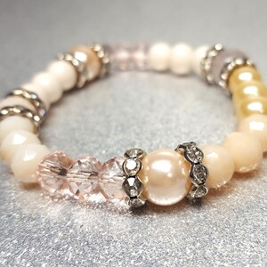 Puede incluir: Un delicado brazalete hecho con cuentas blancas, rosadas y doradas. El brazalete presenta una gran cuenta de perla adornada con diamantes de imitación.