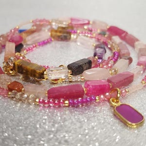 Pode incluir: Colar de várias voltas com pedras preciosas rosa, rosa e transparentes, incluindo contas retangulares e redondas. Um pingente dourado com uma pedra rosa pende do colar. O colar está sobre uma superfície prateada.