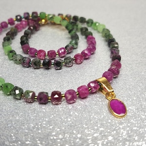 Könnte beinhalten: Ein Halsketten- und Armbandset mit facettierten, würfelförmigen Perlen in abwechselnden Tönen von leuchtendem Pink, tiefem Rot und Waldgrün. Die Halskette enthält eine goldfarbene Metallöse mit einem einzelnen, ovalen, polierten pinken Anhänger.