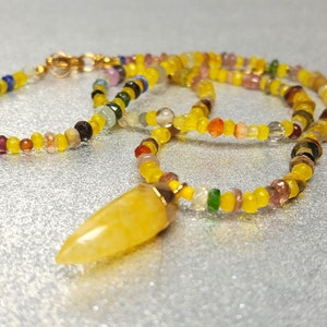 Può includere: Una collana con un ciondolo di pietra gialla e una catena di perline multicolori. Il ciondolo è lungo e appuntito, e la catena è composta da piccole perline rotonde di vari colori, tra cui giallo, rosso, verde e blu.