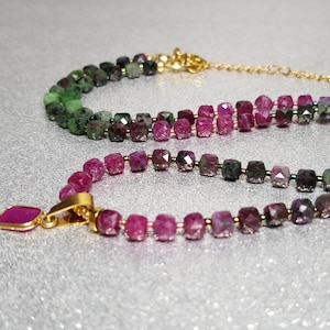 Może przedstawiać: Dwa naszyjniki z koralików ze złotym zapięciem i wisiorkiem. Koraliki przechodzą od zielonego do ciemnofioletowego do różowego. Wisiorek to różowy kamień szlachetny w złotej oprawie. Naszyjniki leżą na srebrnej, błyszczącej powierzchni.