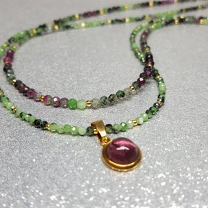 Può includere: Una collana a più fili composta da piccole perline sfaccettate verdi e viola alternate a minuscoli distanziatori color oro. Un ciondolo rotondo a cabochon viola liscio, incastonato in una montatura in metallo color oro, pende dal centro.