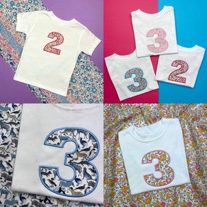 Puede incluir: Una colecci&oacute;n de camisetas blancas de manga corta con grandes n&uacute;meros aplicados en la parte delantera. Los n&uacute;meros est&aacute;n rellenos con varios patrones florales y de animales, incluyendo un n&uacute;mero 2 con flores rosas y un n&uacute;mero 3 con estampados de animales azules.
