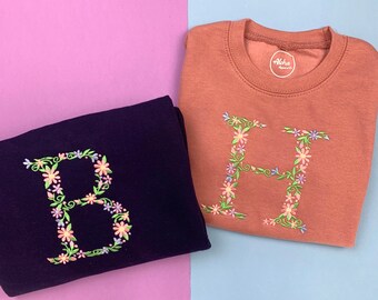 May include: A dark purple sweatshirt and a terracotta sweatshirt, each featuring a large embroidered letter B or H respectively. The letters are decorated with colorful floral and vine patterns. The terracotta sweatshirt has an Aloha Apparel Co label.