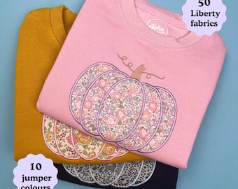 Peut inclure: Trois sweat-shirts à col rond en rose, jaune moutarde et bleu marine. Chacun présente une grande applique de citrouille avec un motif floral Liberty. Le texte indique 50 Liberty fabrics et 10 jumper colours, avec une étiquette Aloha.